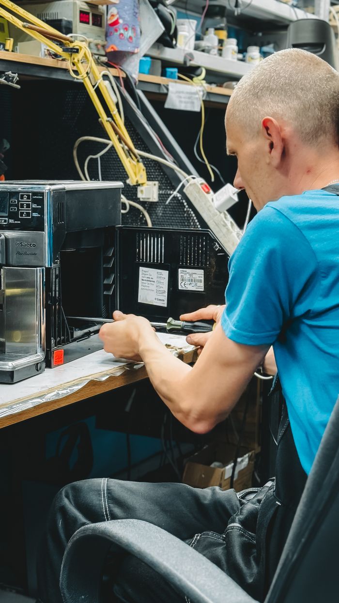 services-01 A skilled technician repairs a coffee machine in a cluttered electronic workshop.